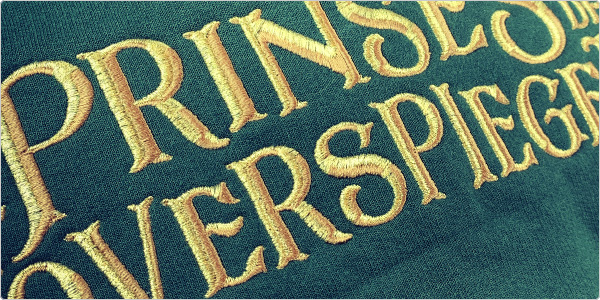 close up van geborduurd logo van De Prinses en de Toverspiegel op een groene hoodie met gouden letters.