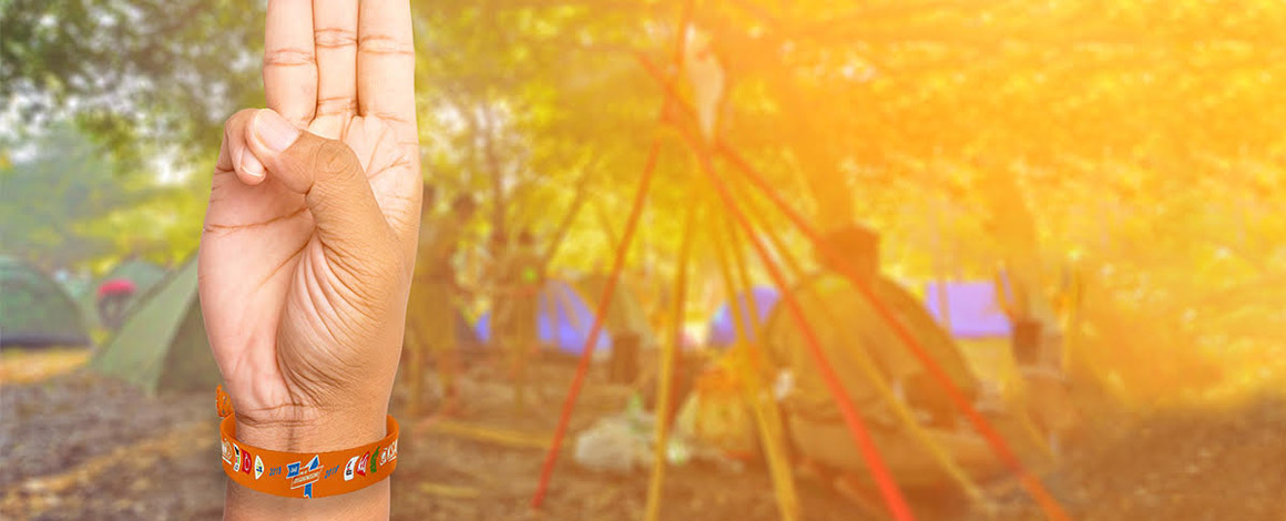 arm met polsbandje aan van Dag van de jeugdbeweging met op de achtergrond een onscherpe afbeelding van een kamp-locatie
