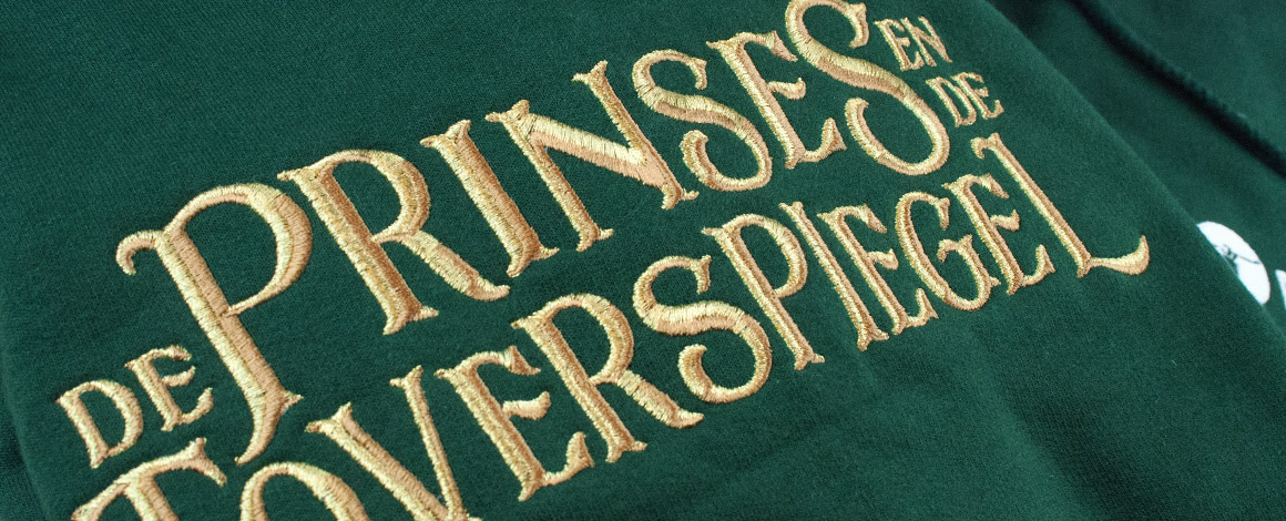 close up van geborduurd logo van De Prinses en de Toverspiegel op een groene hoodie met gouden letters.